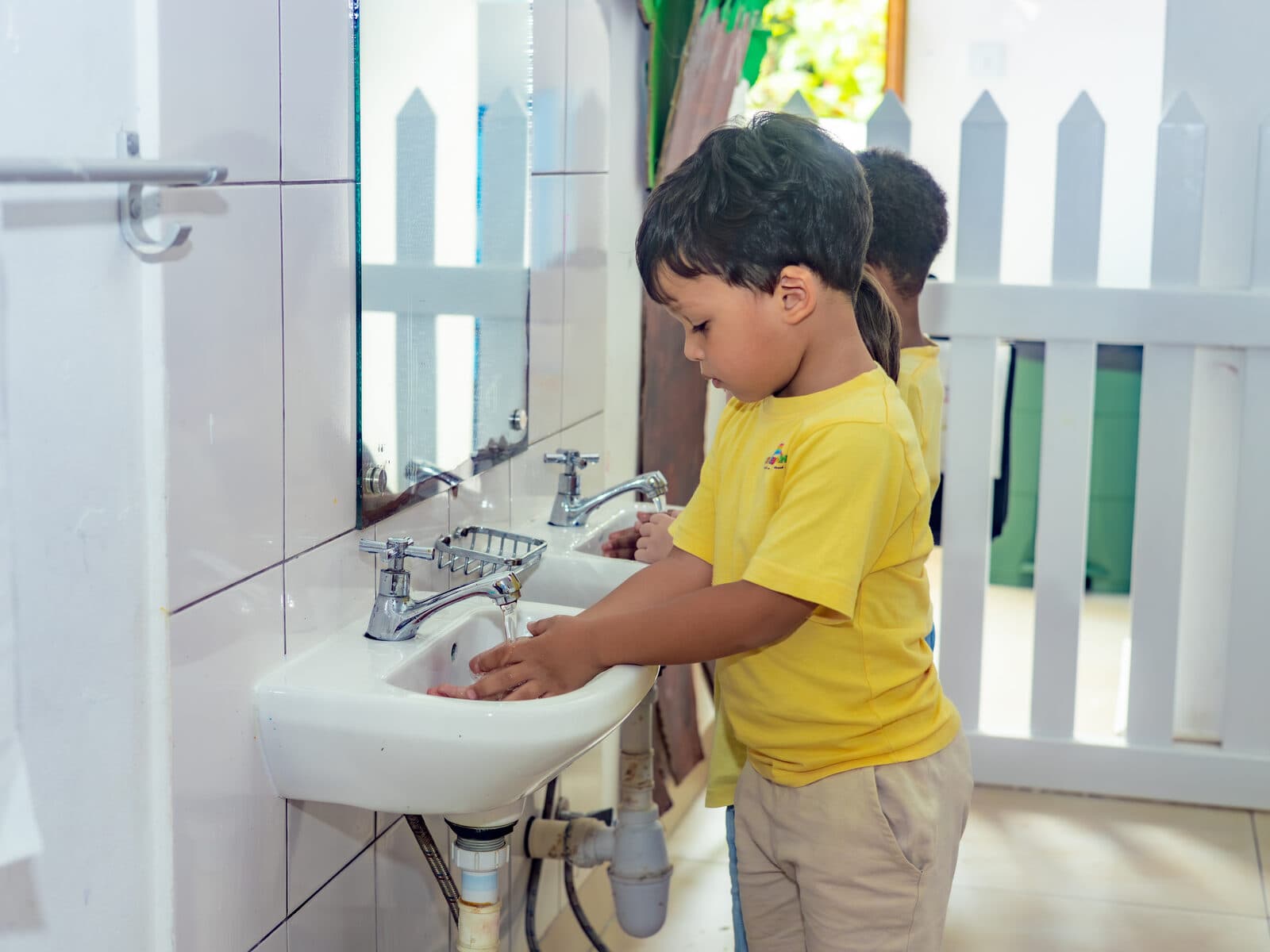 A teacher helping children wash their hands at the classroom sink beneath a wash-your-hands sign