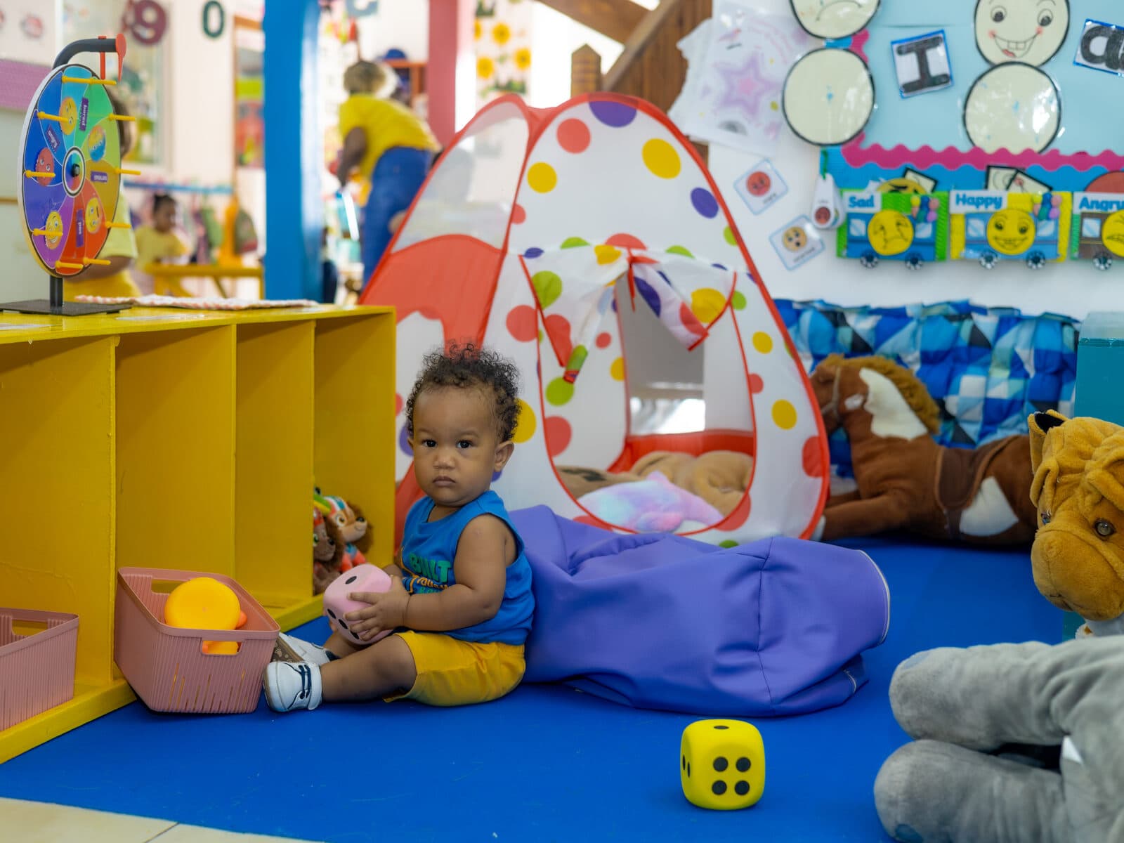 A toddler sitting by a play tent in a bright, colourful indoor play area